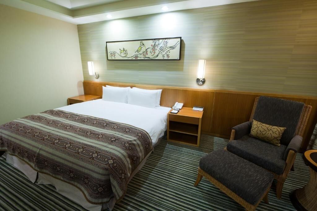 A hotel room with a large bed, a wooden headboard, a chair, a nightstand with a phone, and a wall-mounted artwork.