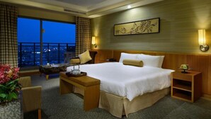 Habitación de hotel con una cama grande, un escritorio, una silla, una mesita de noche y vistas de la ciudad de noche.