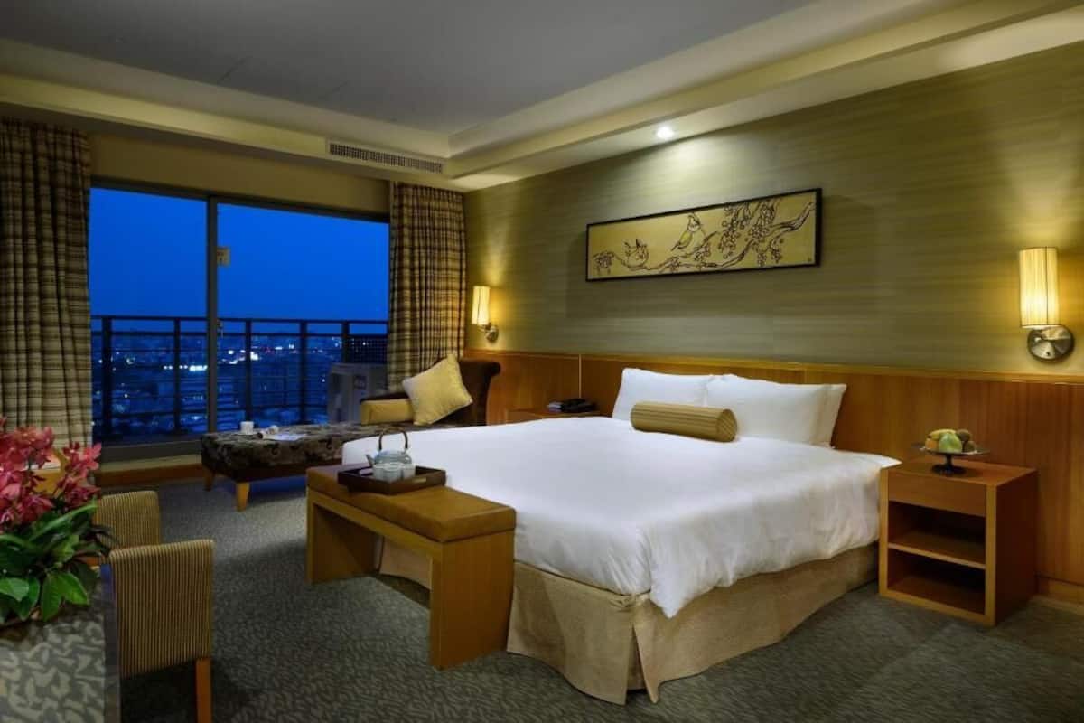 Habitación de hotel con una cama grande, un escritorio, una silla, una mesita de noche y vistas de la ciudad de noche.
