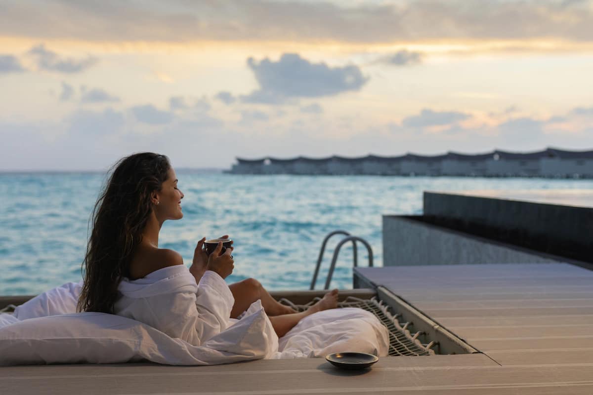 A person relaxing on a sun lounger by the sea, holding a cup, with a view of the ocean and distant overwater structures.