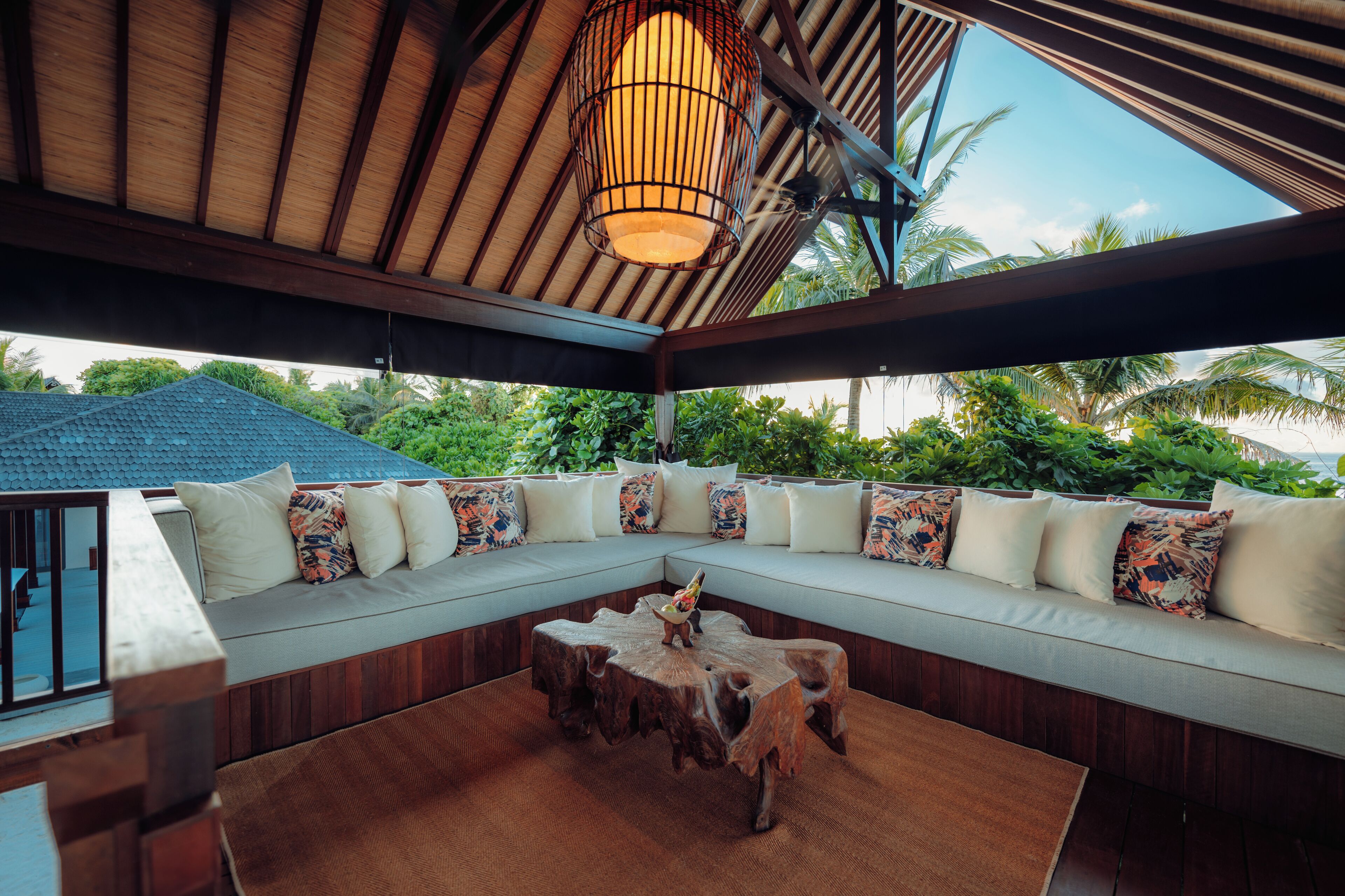 A covered outdoor seating area with a wooden ceiling, a woven light fixture, and a view of lush greenery.