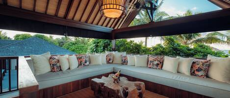 A covered outdoor seating area with a wooden ceiling, a woven light fixture, and a view of lush greenery.