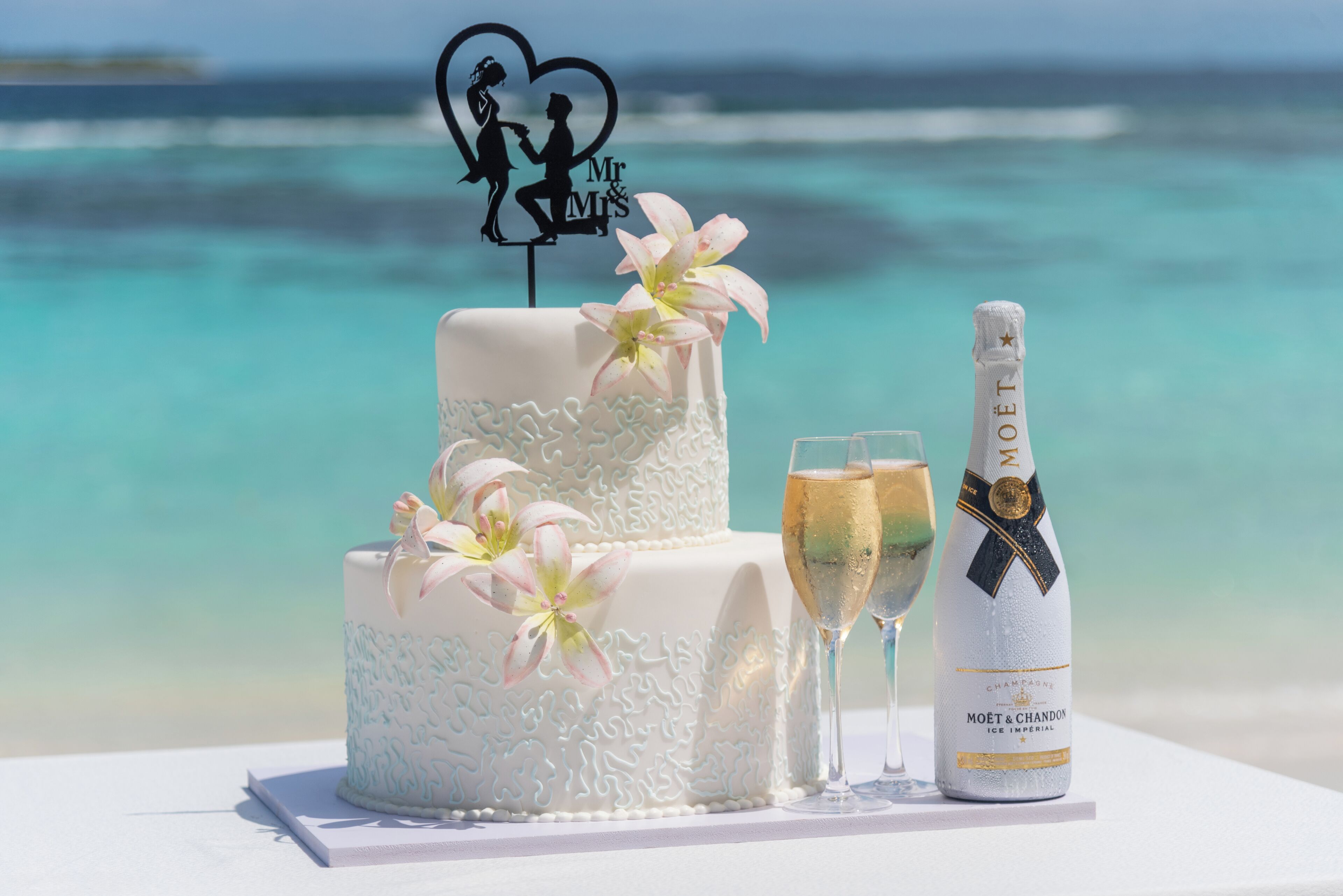 Un gâteau de mariage à deux étages, surmonté d’un décor en forme de cœur et orné de fleurs, est placé à côté de deux coupes à champagne et d’une bouteille de champagne Moët & Chandon Ice Imperial sur une table offrant une vue sur l’océan.