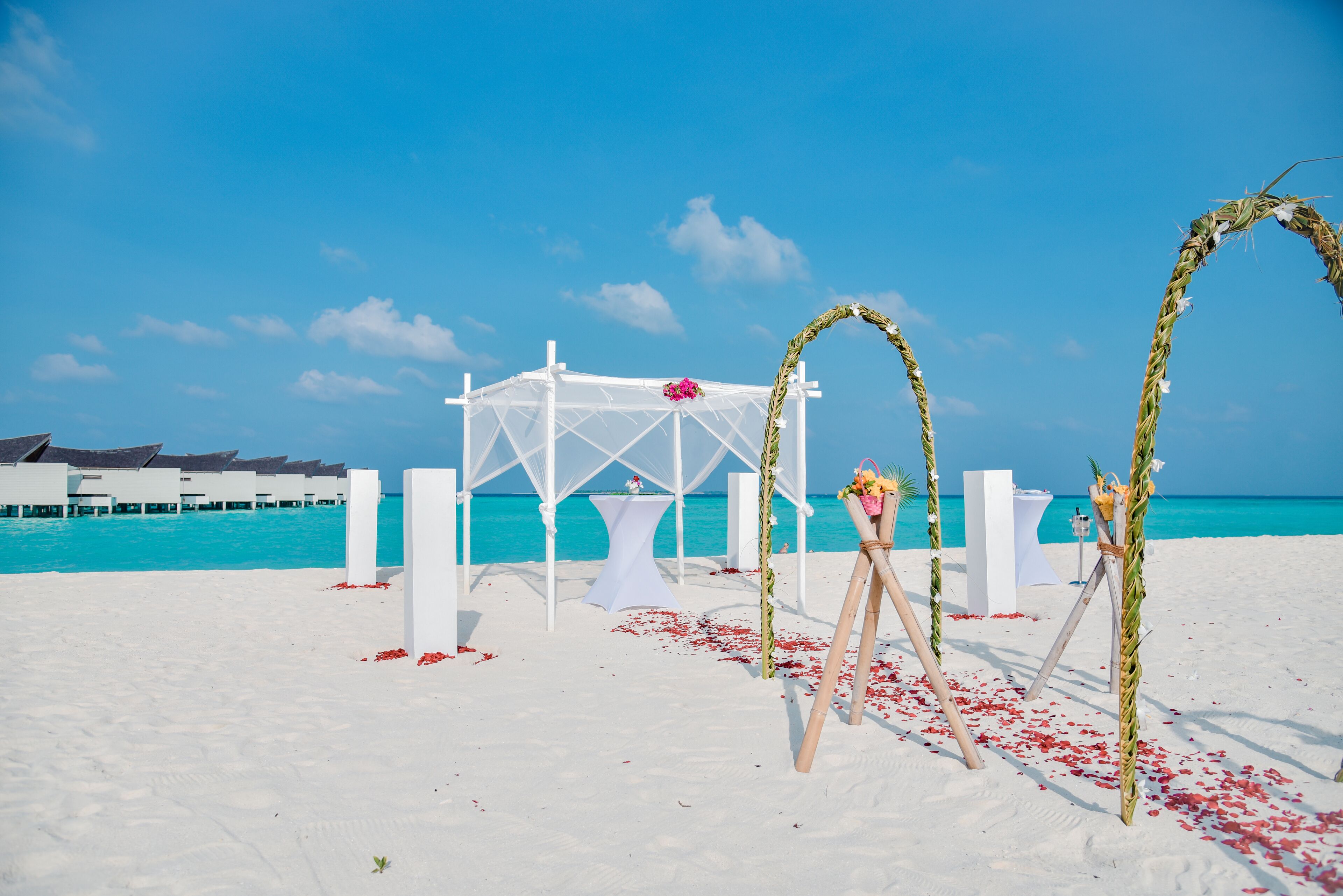 A beach wedding setup with a white archway, a small table with a flower arrangement, and a backdrop of turquoise water.