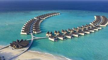 Luchtbeeld van waterwoningen gerangschikt in een halve cirkel, verbonden door looppaden, met een strand en palmbomen op de voorgrond.