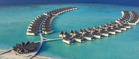 An aerial view of overwater bungalows arranged in a semi-circular pattern, connected by walkways, with a beach and palm trees in the foreground.