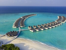 Vista aérea de bungalows sobre el agua dispuestos en un patrón semicircular, conectados por pasarelas, con una playa y palmeras en primer plano.