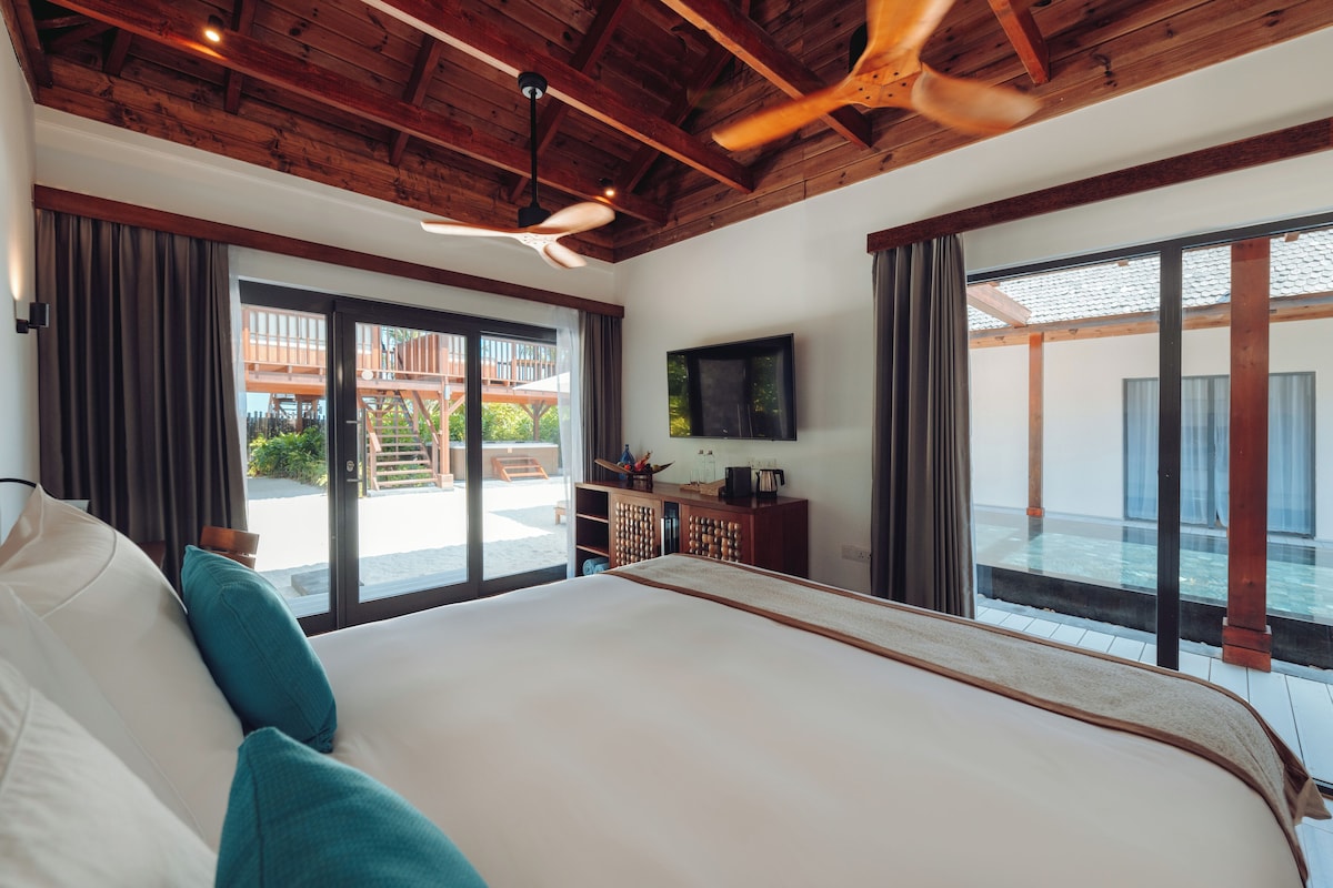 Een ruime slaapkamer met een groot bed, houten balken in het plafond, een plafondventilator en een schuifdeur van glas die uitkomt op een buitenruimte.