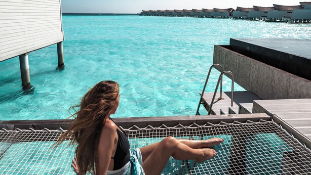 Een vrouw zit aan het zwembad met uitzicht op de oceaan en bungalows op palen.