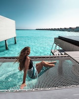 Una mujer sentada junto a la piscina con vistas al océano y bungalows sobre el agua.