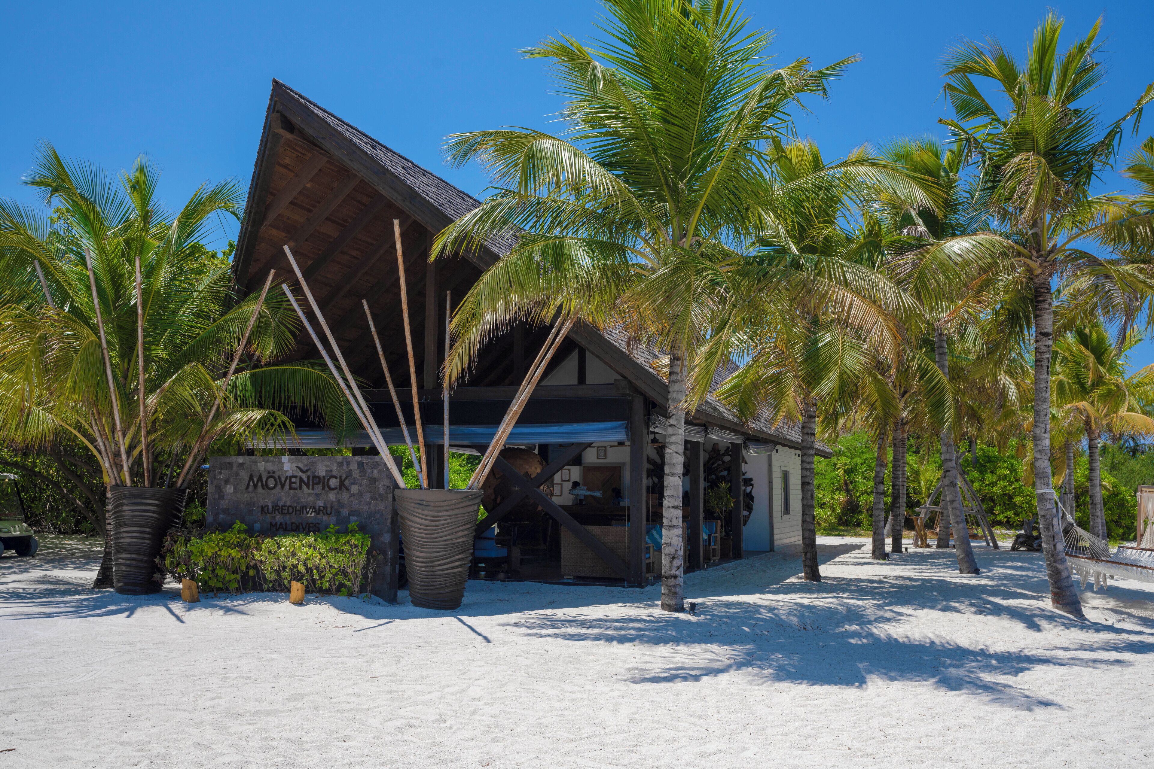 A beachside building with a sloped roof, surrounded by palm trees and a sign reading "Mövenpick Kurehdivaru Maldives".