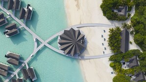 Vista aérea de un resort con una estructura circular central, conectada por un puente a una playa y una zona acuática con edificios.