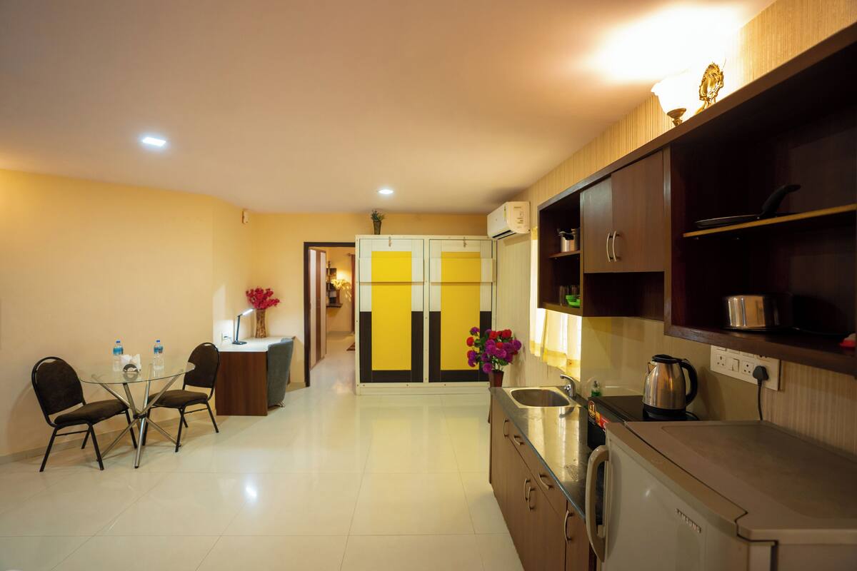 A modern kitchen with a built-in oven, a sink, and a countertop. There is a dining area with a glass table and chairs. The kitchen has wooden cabinets and a yellow and black door.