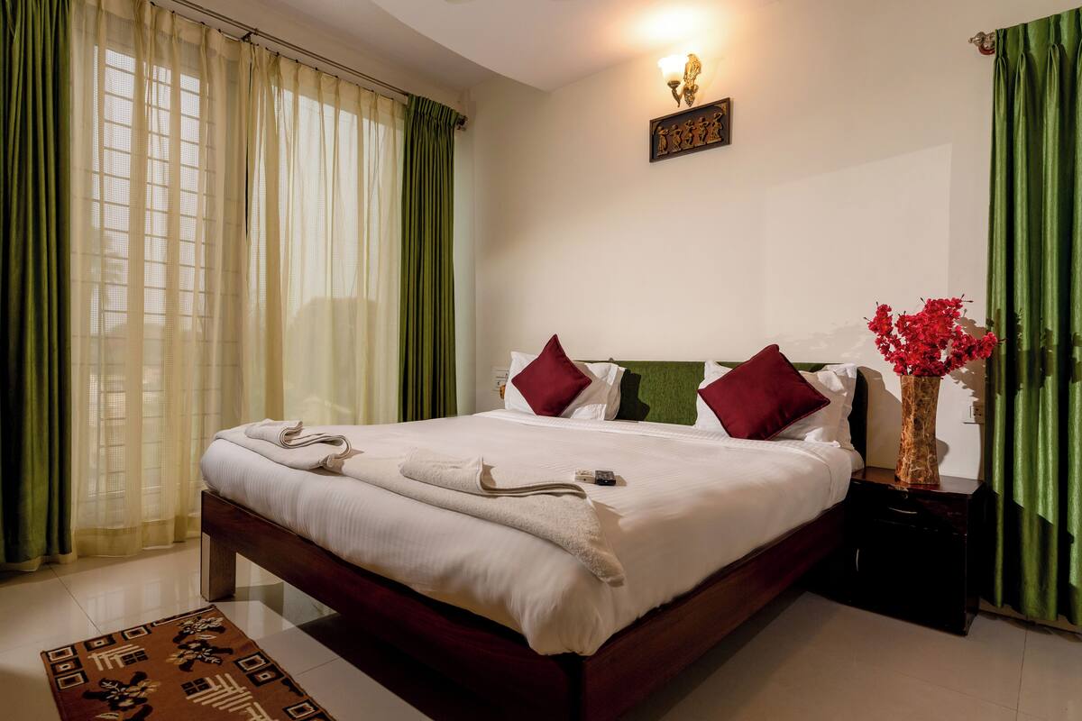 A neatly made bed with white linens, a red vase with red flowers, and a small nightstand with a lamp.