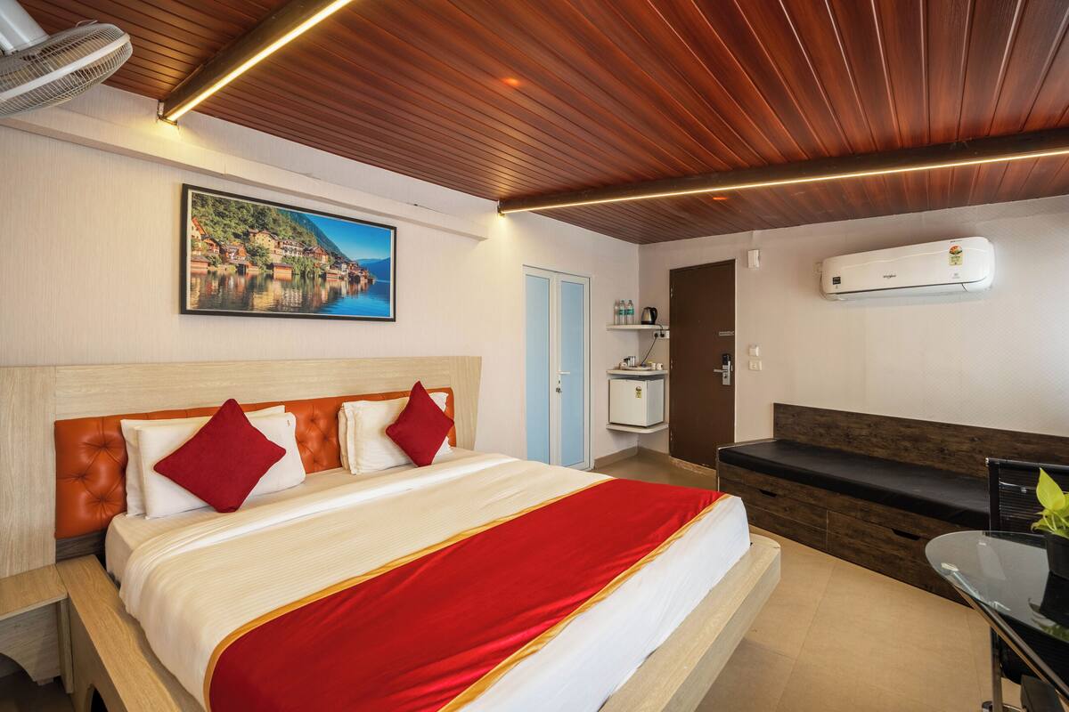 A hotel room with a large bed, a wooden ceiling, an air conditioner, a painting, a bedside table, and a bench.