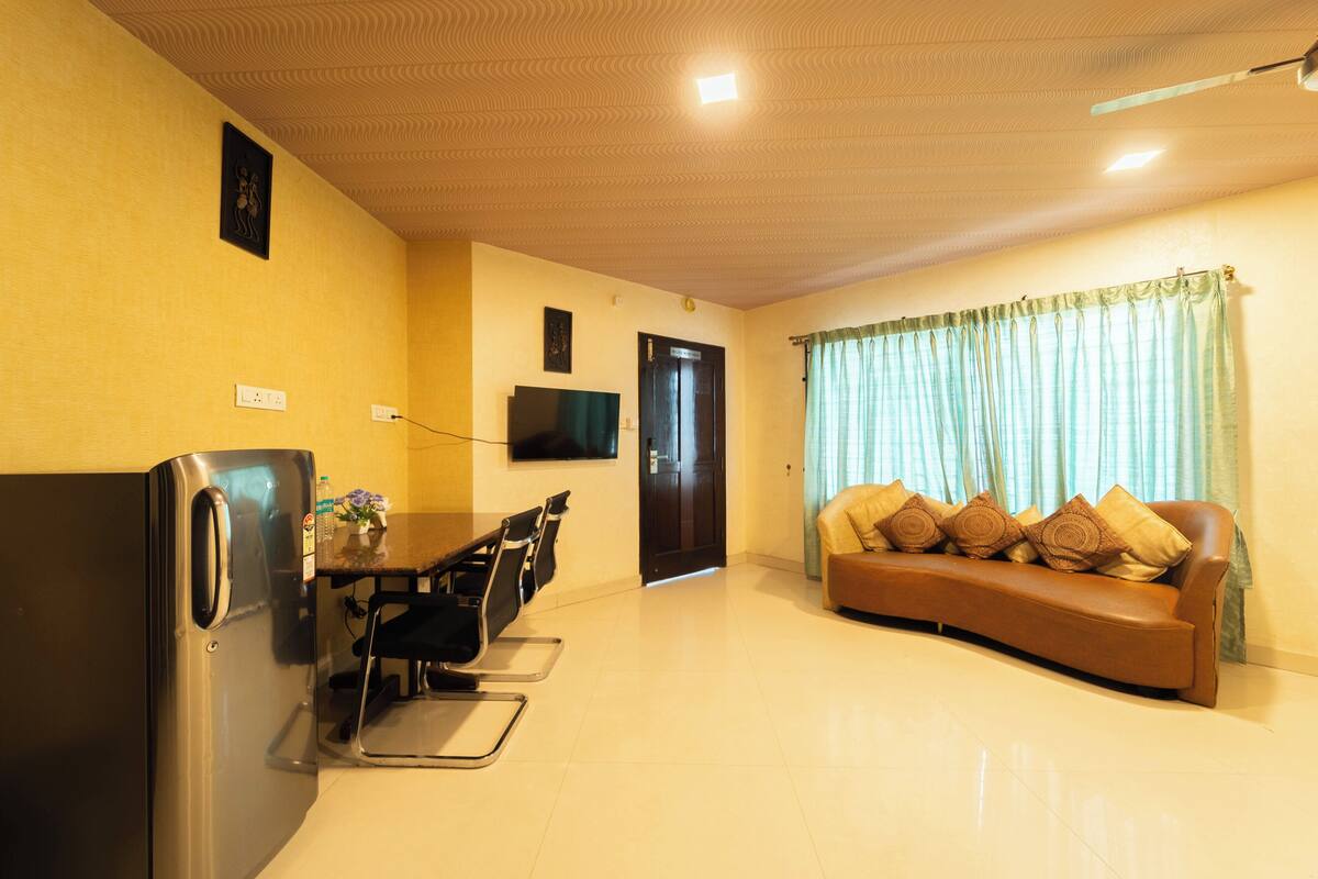 A living room with a sofa, a dining table with chairs, a refrigerator, and a television mounted on the wall.
