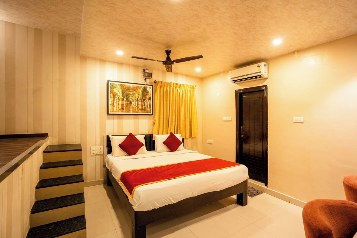 A hotel room with a bed, a ceiling fan, an air conditioning unit, a painting on the wall, and a staircase.