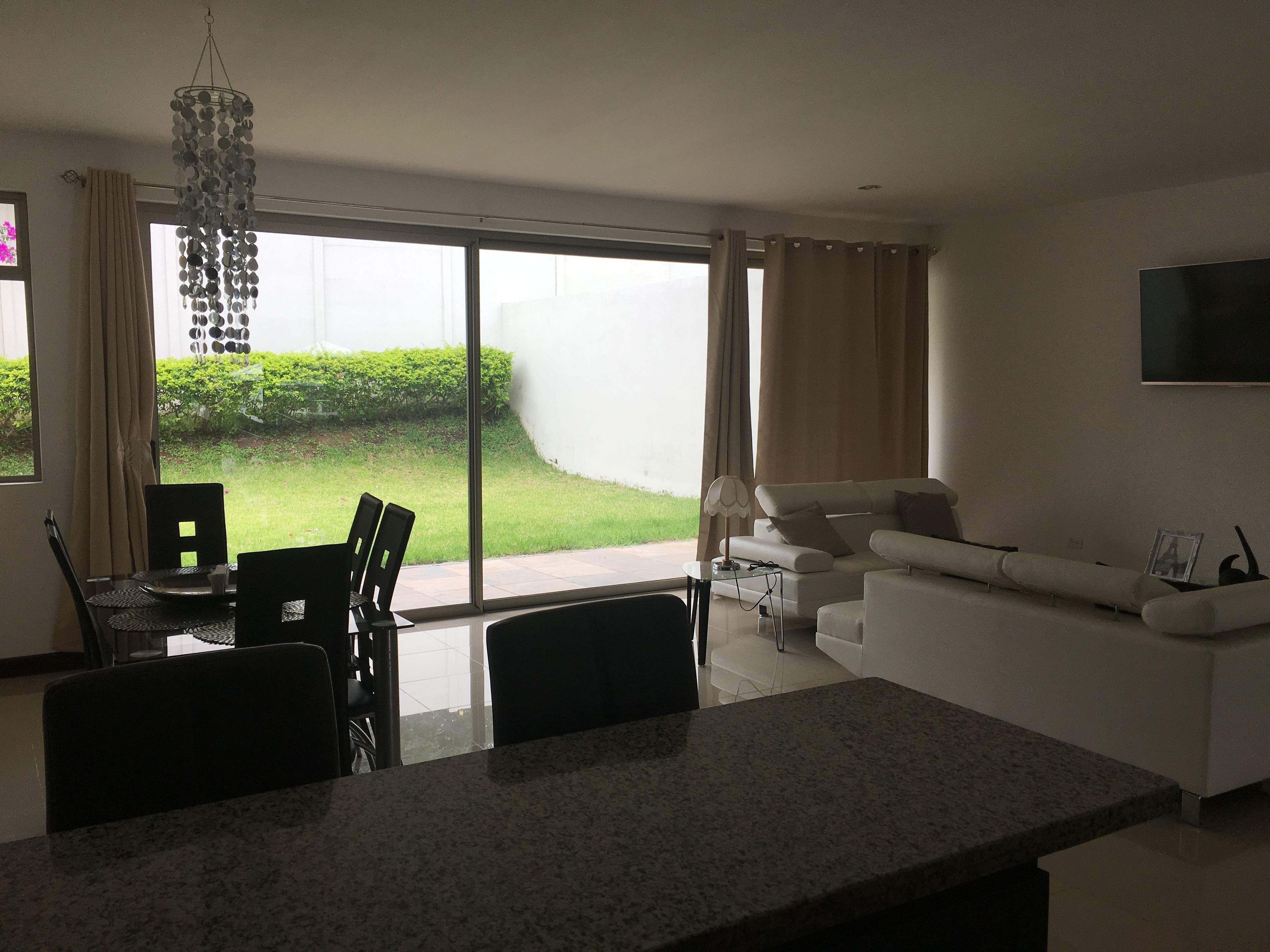 Ett modernt vardagsrum med ett stort fönster, en matplats med stolar, en soffa och en platt-TV som är monterad på väggen.