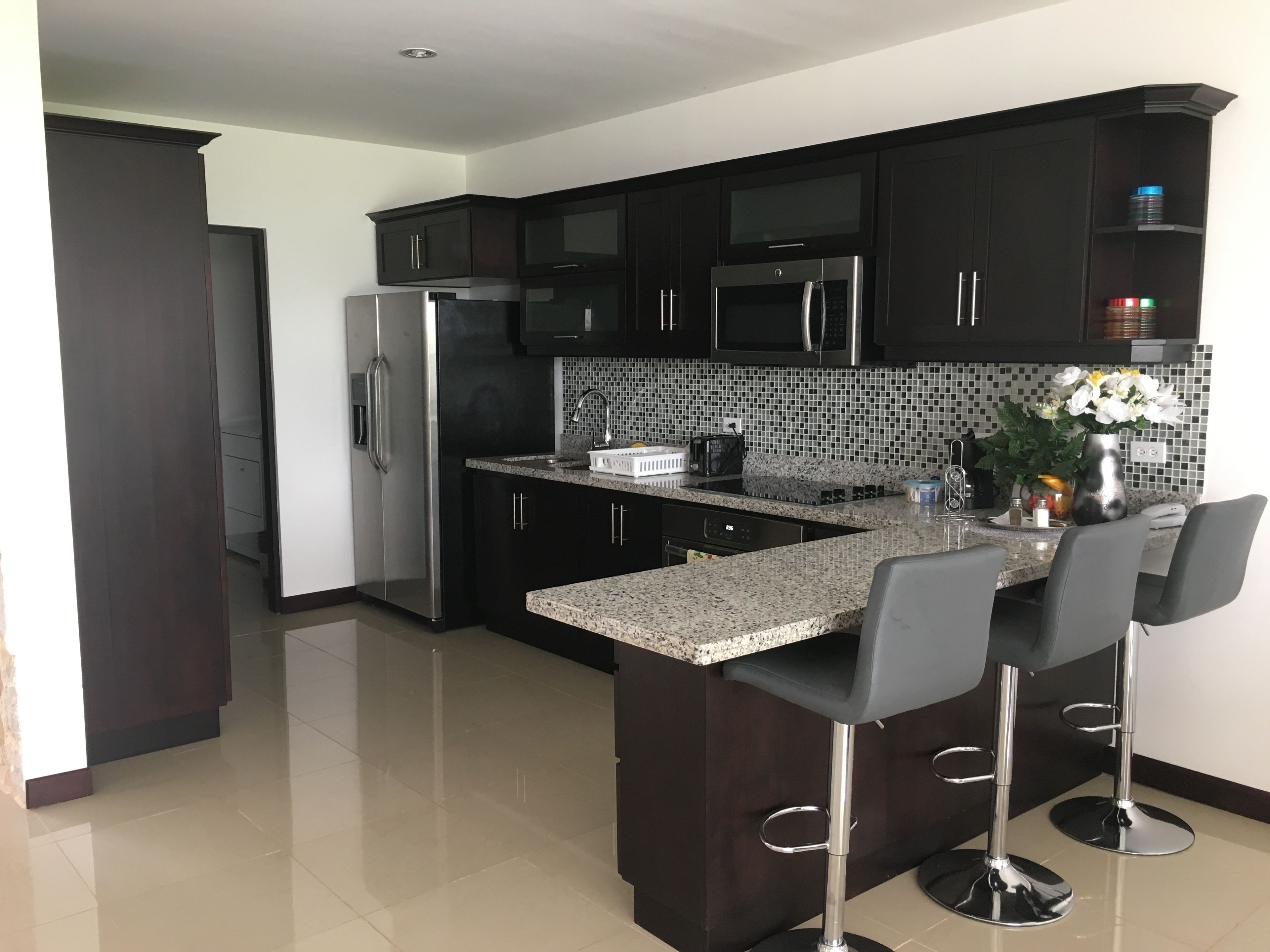 A modern kitchen with dark cabinetry, stainless steel appliances, a granite countertop, and a bar area with stools.