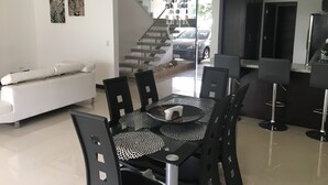 A modern dining area with a glass table, black chairs, and a white sofa. A staircase leads to a garage with a car parked outside. - Las Yagrumas Condo Escazu (San Rafael)