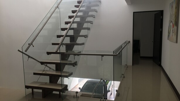 A modern staircase with glass railings and a metal handrail, leading to a landing with a view of a car outside. - Las Yagrumas Condo Escazu (San Rafael)