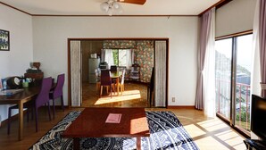 A spacious living area with a dining table, chairs, and a television. There is a ceiling fan, a large window with curtains, and a view of the outside. - Japan Countryside Atami (Atami)