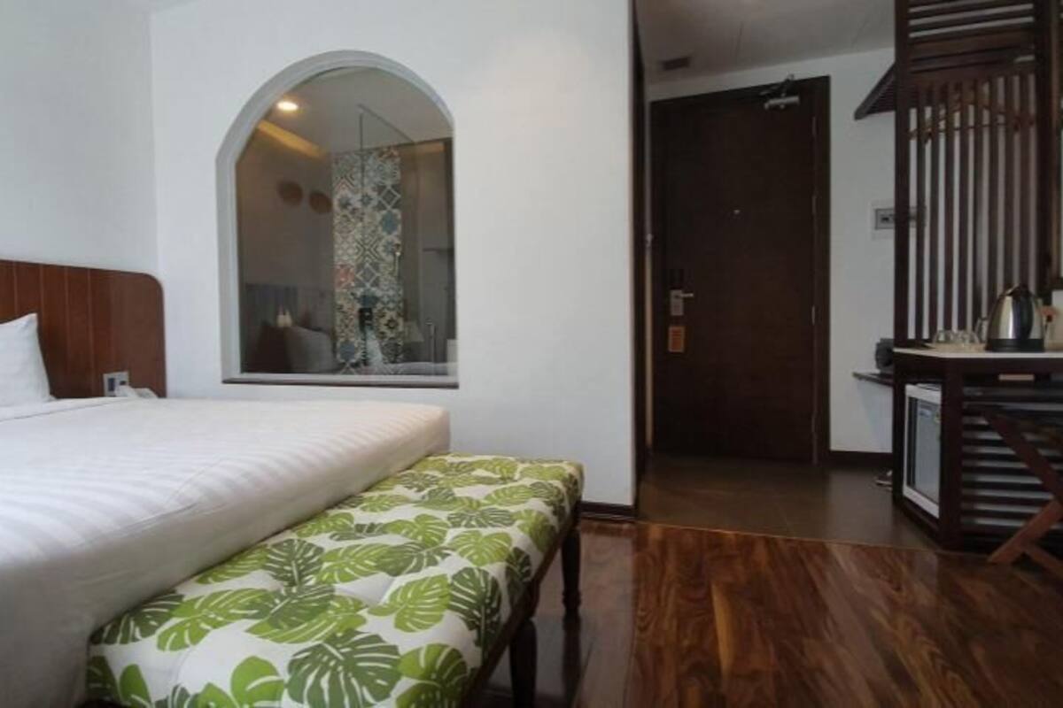 Habitación de hotel con una cama grande, un banco, piso de madera, un espejo y una puerta que conduce a otra habitación.