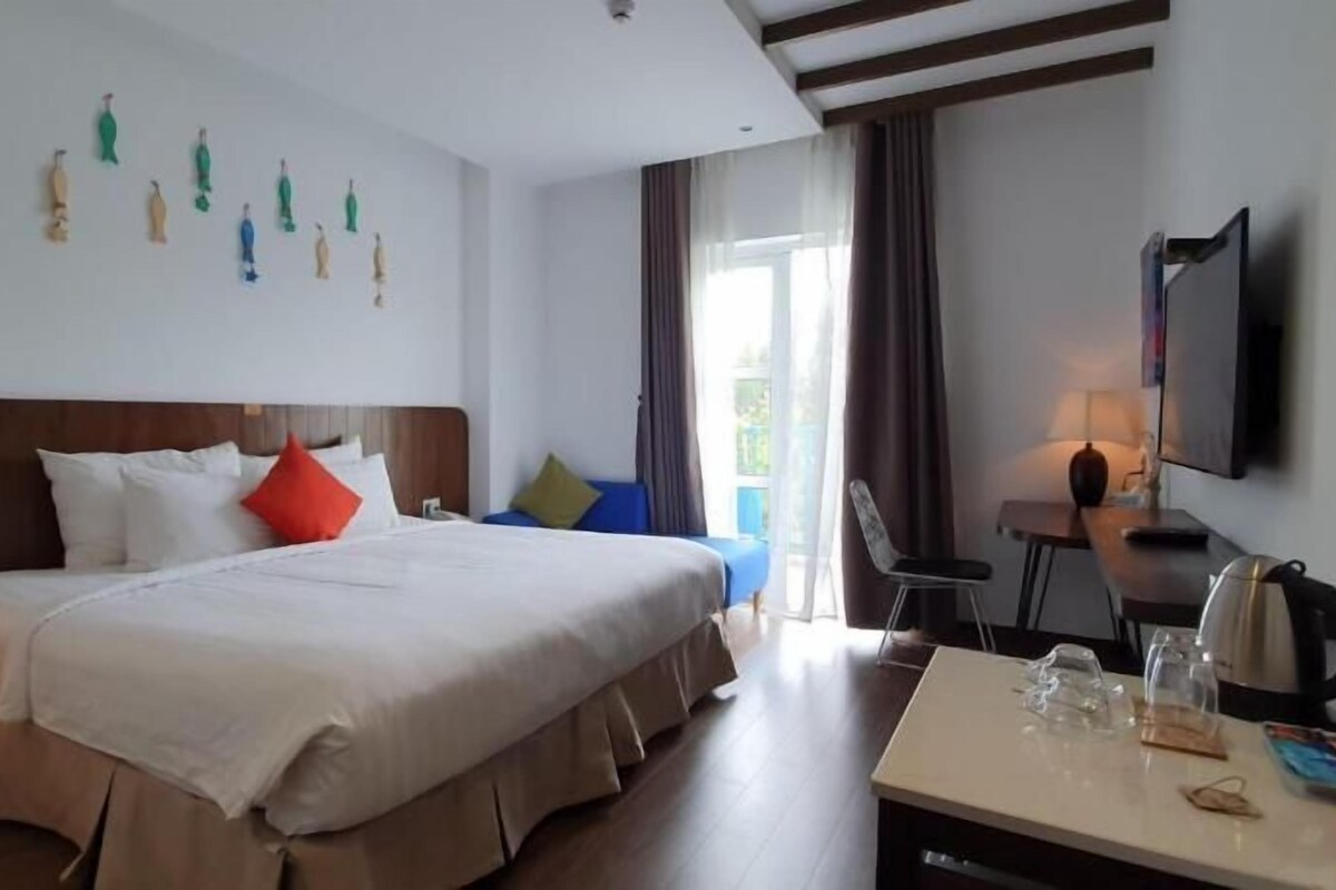A hotel room with a large bed, a desk with a kettle, a chair, and a TV.