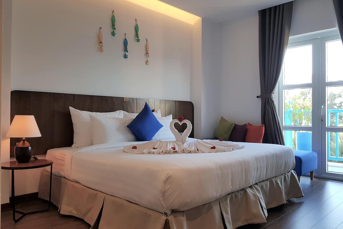 Una cama bien hecha con una toalla dispuesta en forma de corazón, una lámpara de noche y vistas al exterior a través de un ventanal grande.