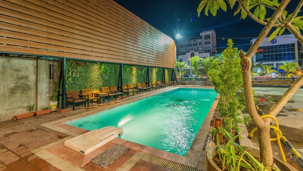 A swimming pool with a wooden deck and lounge chairs, surrounded by greenery and a modern building in the background. - Ruby Myanmar Hotel (Mandalay)