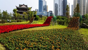A park with flower beds, a traditional building, and modern high-rise buildings in the background.