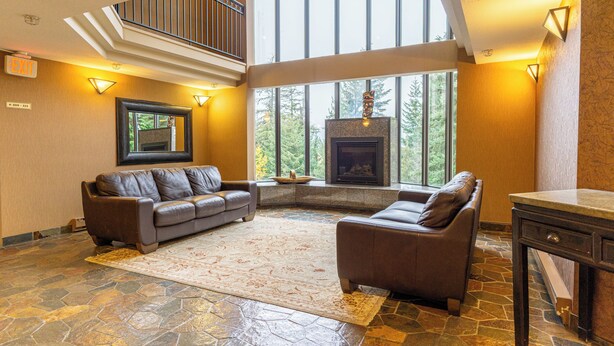 A spacious living room with a stone floor, leather sofas, a fireplace, and large windows offering a view of trees.
