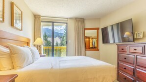 A hotel room with a large bed, a wooden dresser, a flat-screen TV, and a balcony with a mountain view.