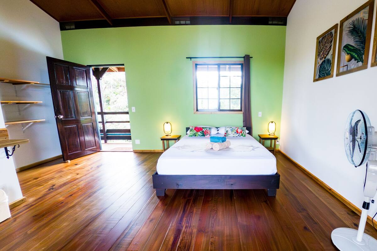 Una habitación con una cama, piso de madera, una ventana y un ventilador.