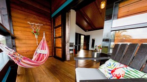 A room with wooden floors, a hammock, a bed with a colorful pillow, and a view of the outside. - El Caribeo - Nature Lodge (Tierra Oscura)