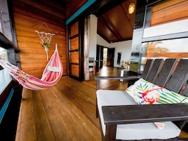 Quarto com piso de madeira, uma rede, uma cama com travesseiro colorido e vista para o exterior.