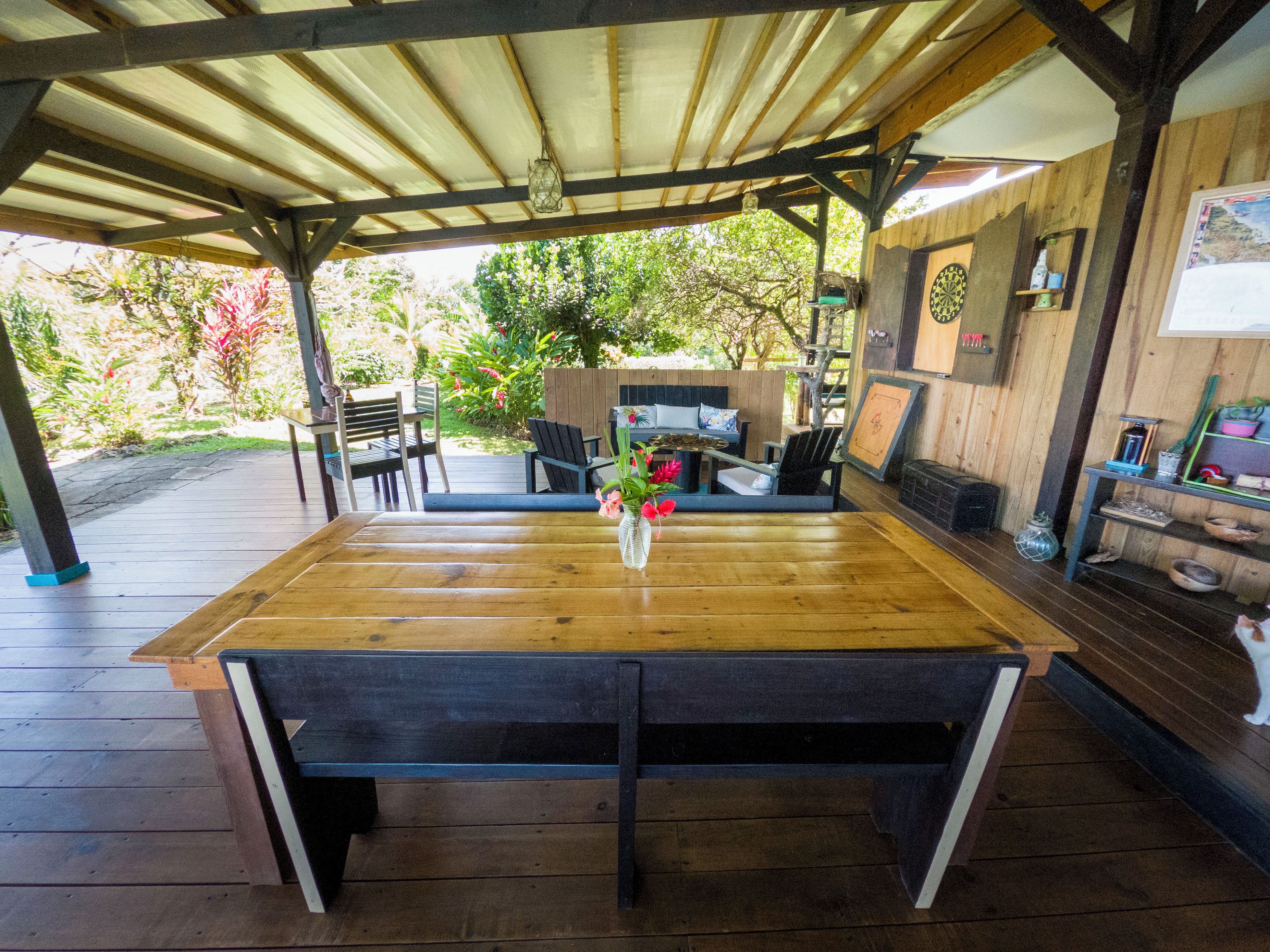 Eine überdachte Terrasse mit Holztisch und -stühlen, durch die offenen Seiten ist ein kleiner Garten sichtbar, und ein rustikales Interieur mit Holzbalken und Regalen.