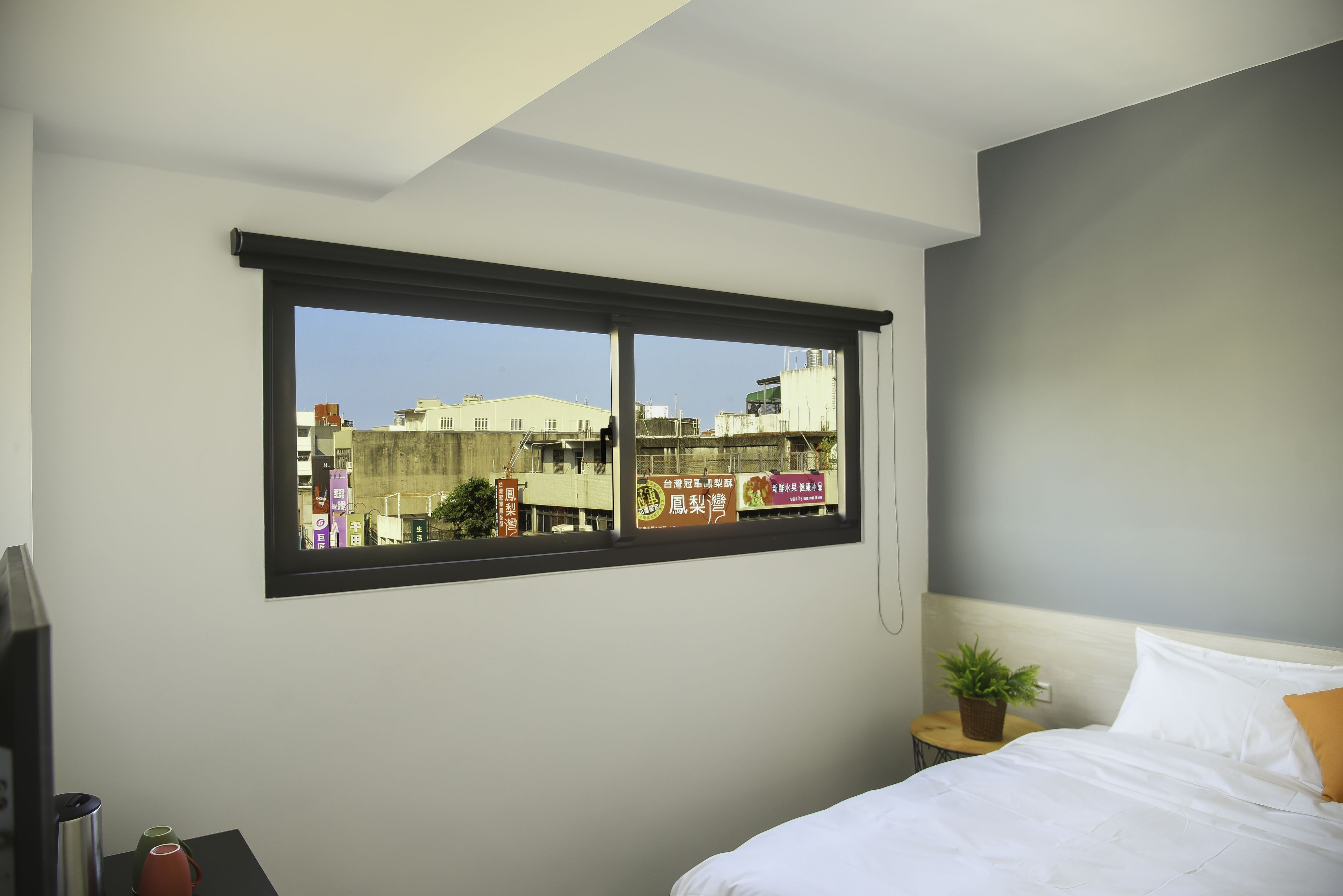 A bedroom with a large window, a bed, a bedside table with a plant, and a TV on a stand.