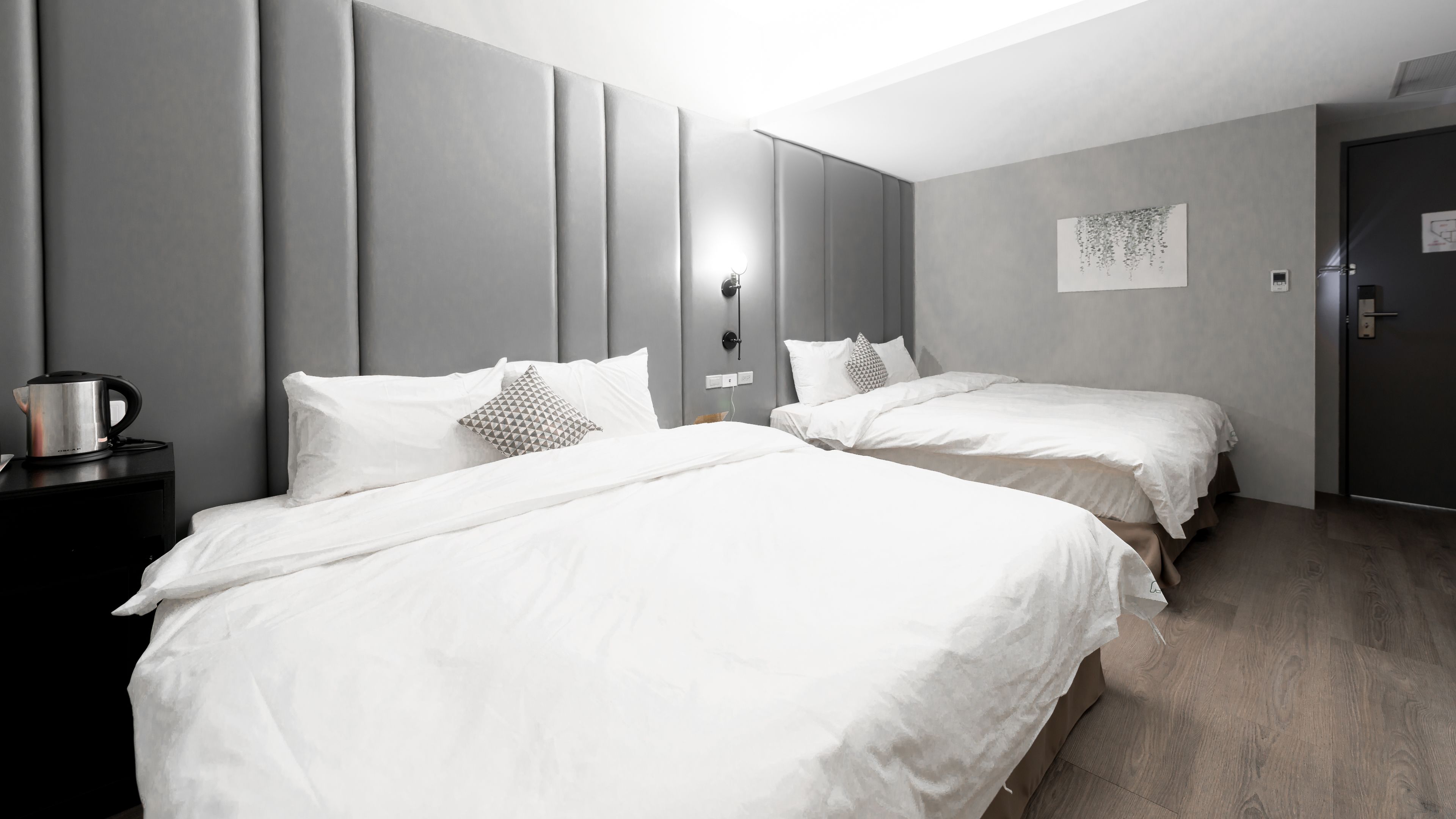 A hotel room with two beds, a bedside table with a kettle, and a wall-mounted light.