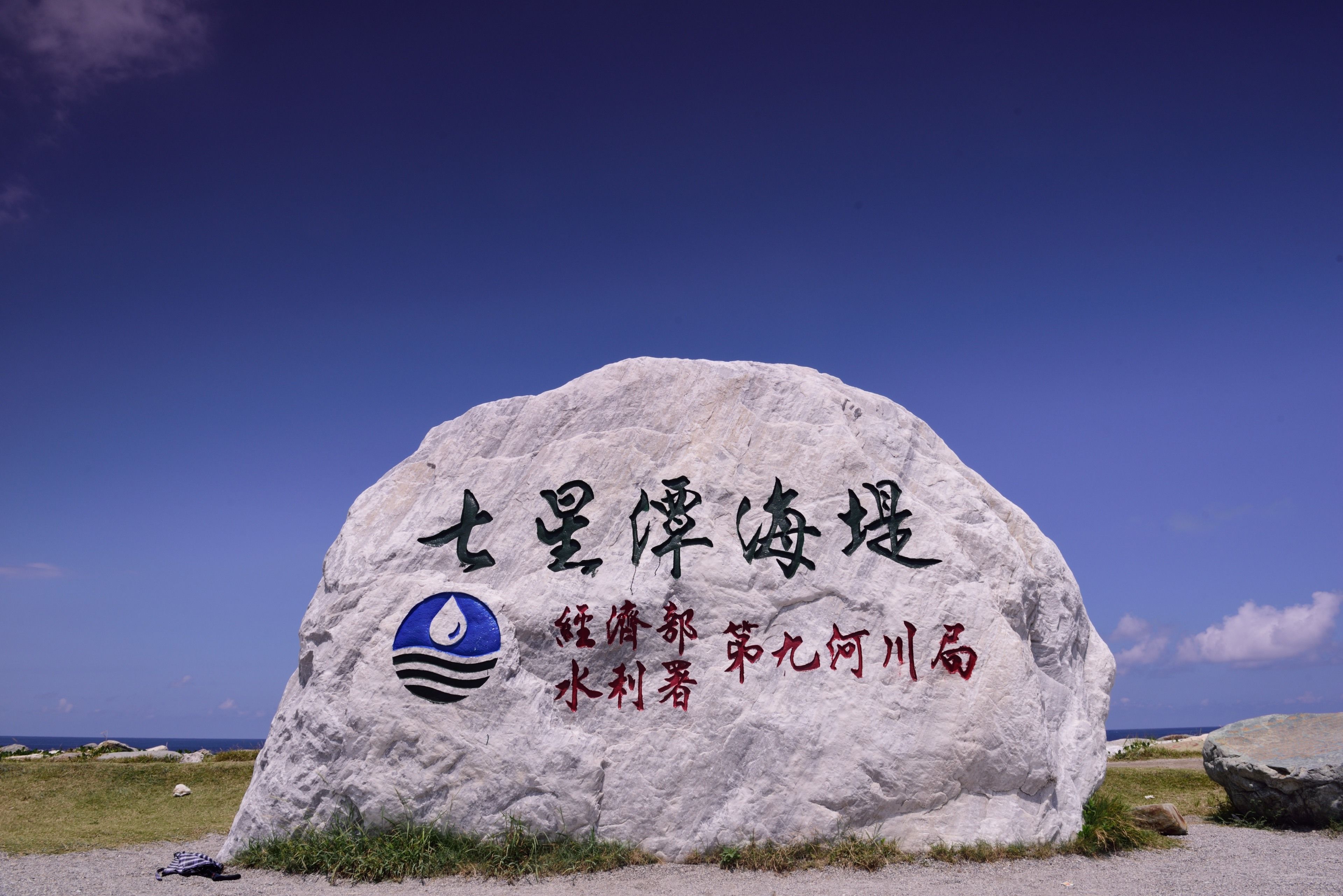 A large rock with inscriptions and a logo, surrounded by a grassy area and the ocean in the background.