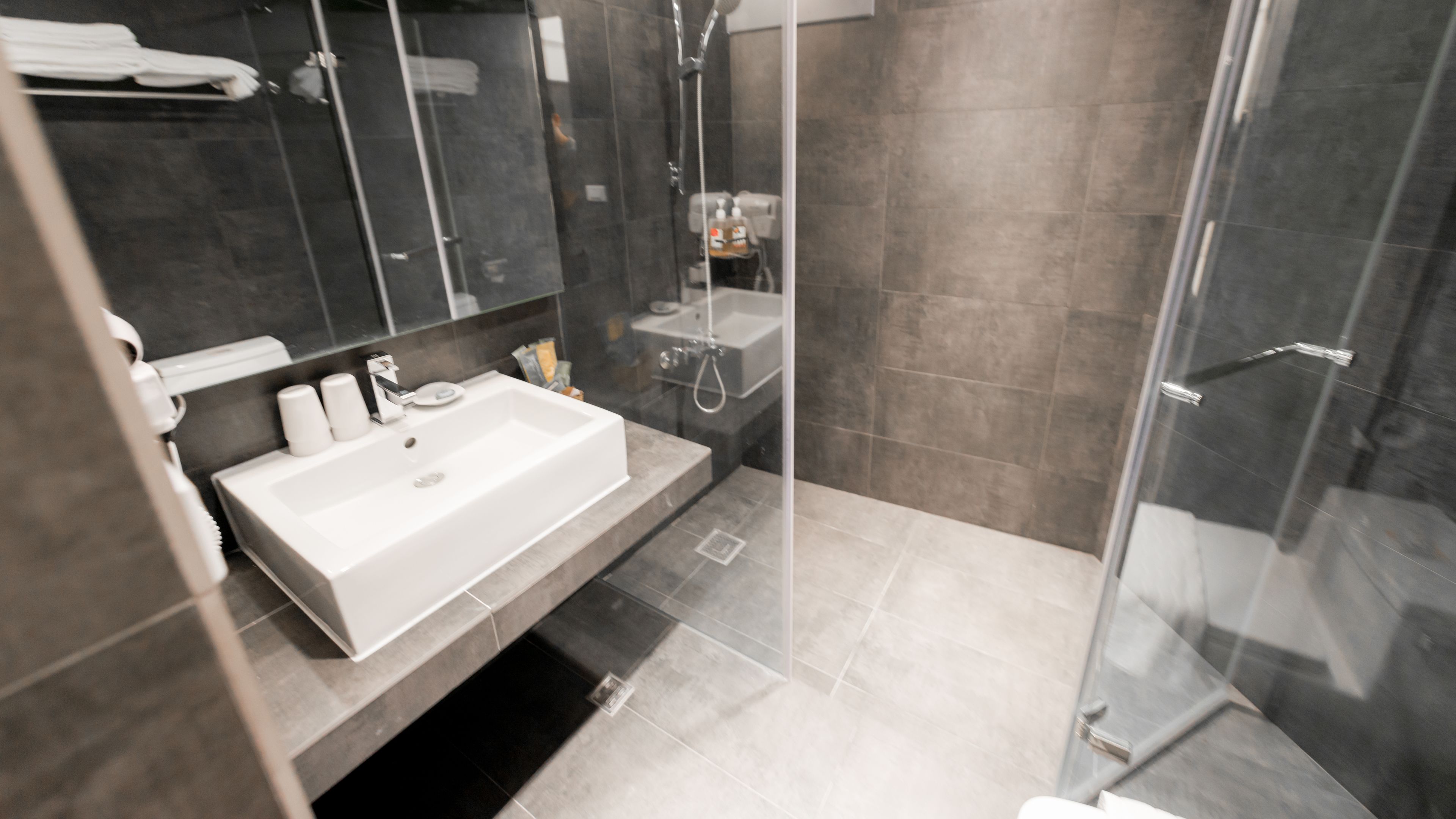 A modern bathroom with a large white sink, a glass shower enclosure, and dark tiled walls.