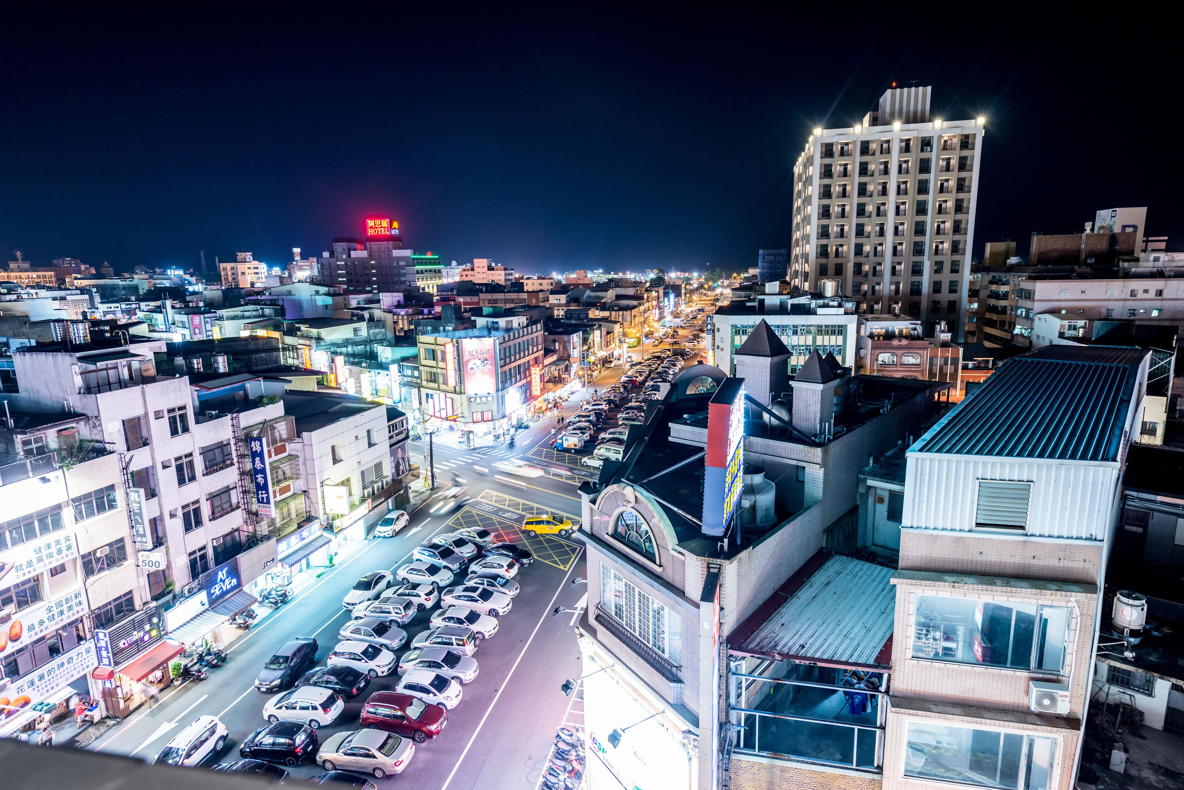 A cityscape at night with a prominent hotel building, a busy street with parked cars, and various commercial buildings.