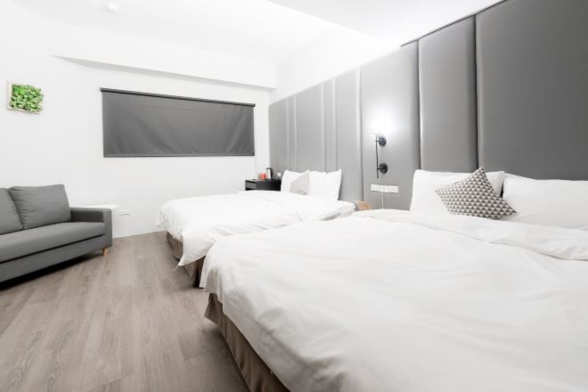 A modern hotel room with two beds, a grey sofa, a small table, and a window with a blind.