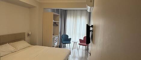 Une chambre d’hôtel avec un grand lit, un bureau, deux chaises, une armoire et une fenêtre avec des rideaux.