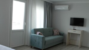 A room with a teal armchair, a small bedside table with a TV, and a window with curtains. - Butik Akvaryum (Adalar)