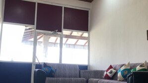 Una sala de estar con techo de madera, un ventilador de techo, un ventanal con persianas, un sofá a rayas y varios cojines.