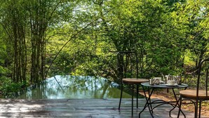 A wooden deck with two metal tables and chairs, overlooking a pond surrounded by lush greenery. - Red Earth Tadoba (Chandrapur)