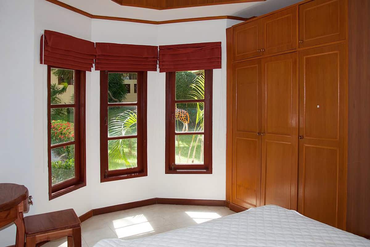 A bedroom with a large bed, a wooden wardrobe, and three windows with red valances.