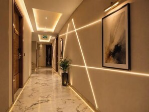 A hotel corridor with a marble floor, framed artwork on the walls, and a potted plant. - Staybook Hotel Nitya Maharani (New Delhi)