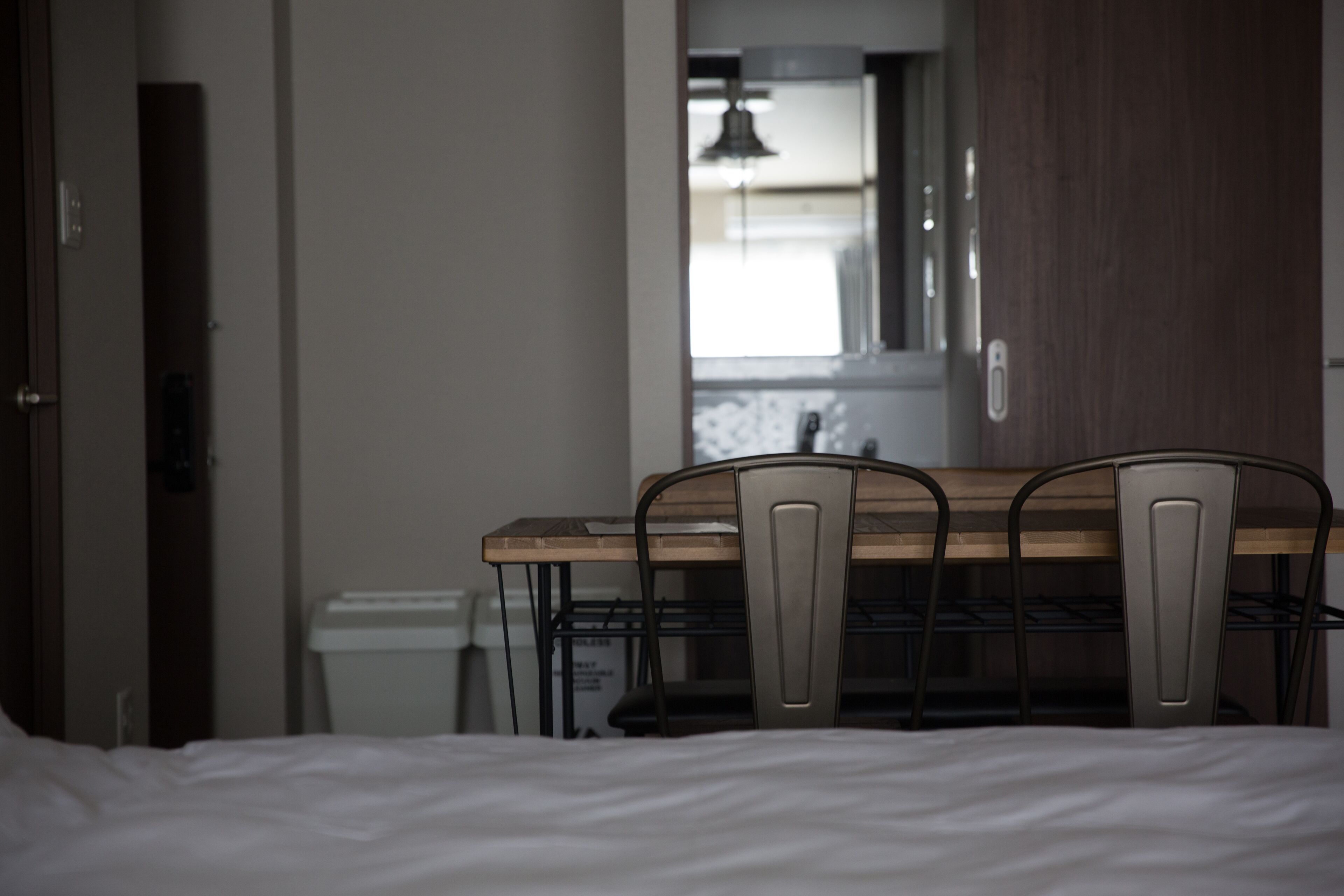 A hotel room with a bed, a wooden table with metal chairs, a small trash bin, and a mirror on the wall.